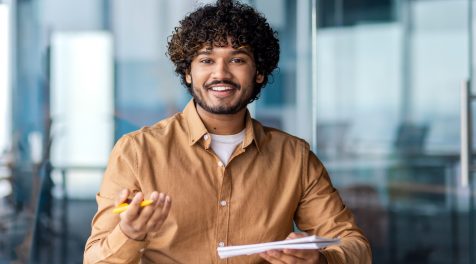Smiling employee sitting in the office conducting a video call interview, holding a notebook in his hands, looking at the webcam.
